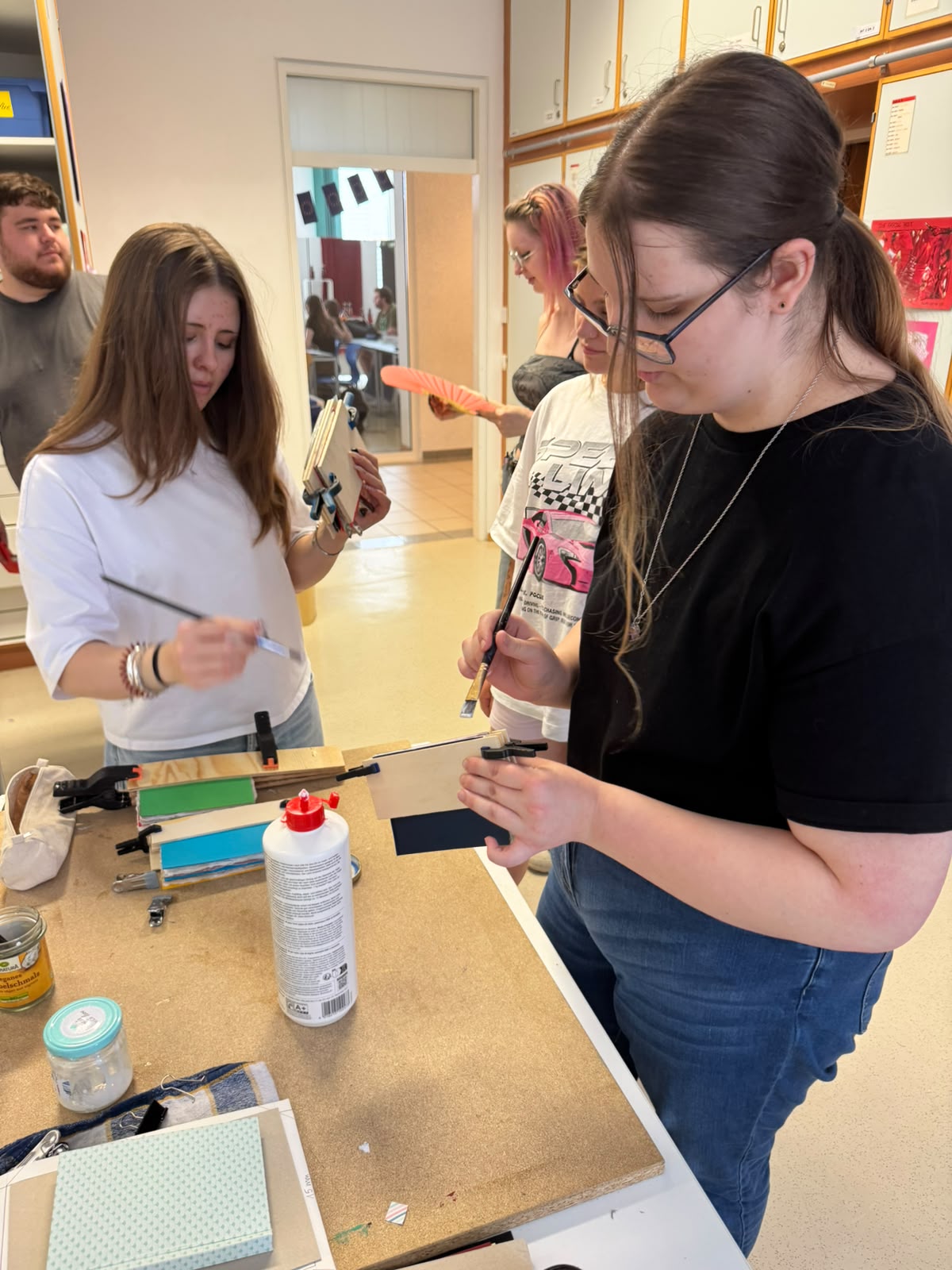 Workshop "Buchbinden" Zwei junge Frauen stehen um einen Tisch und halten Pinsel und Büttenpapier. Eine weiße Leimflasche und andere Utensilien stehen auf der Arbeitsfläche. Im Hintergrund sind weiter Jugendliche zu sehen.
