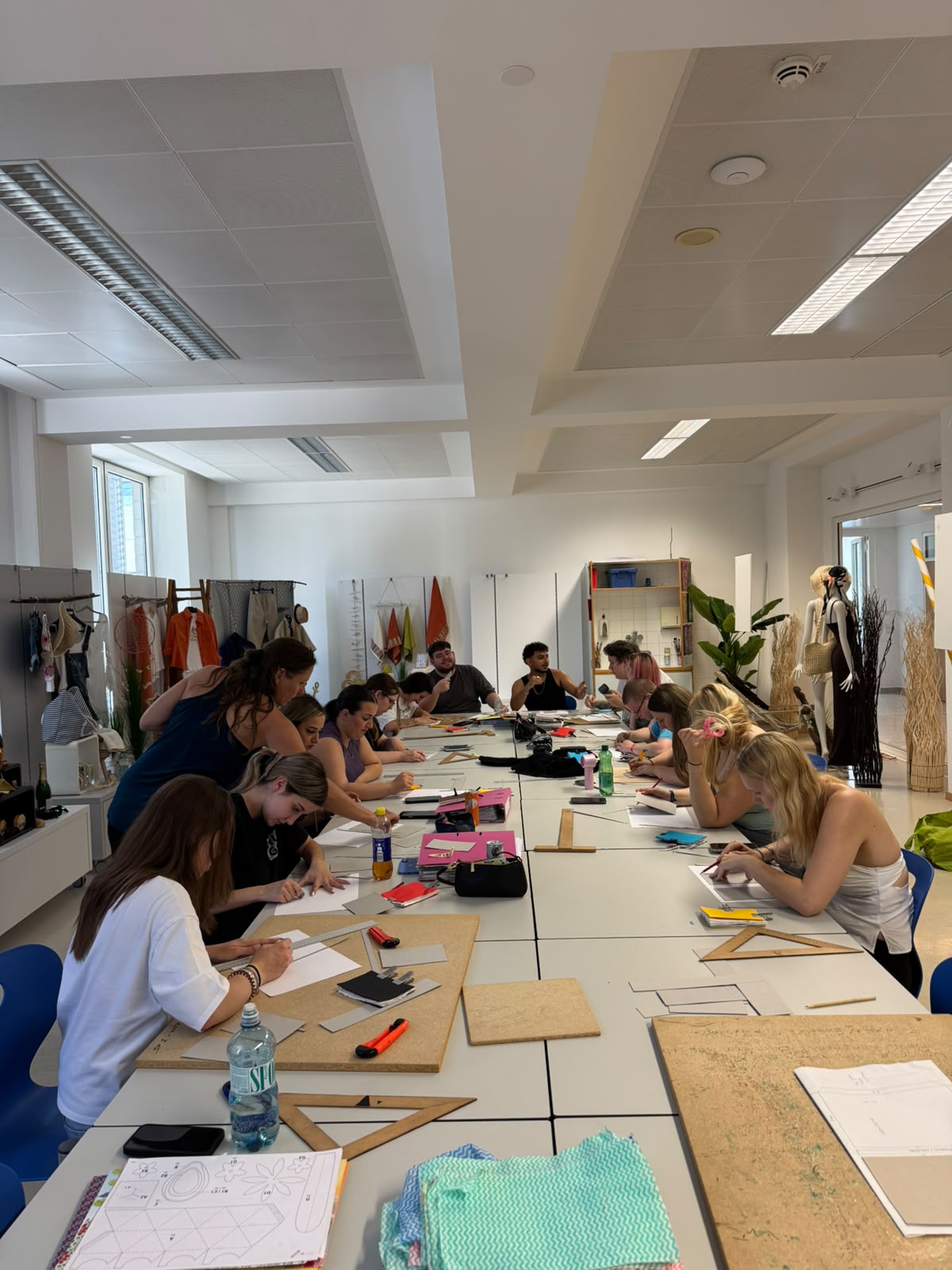 Workshop "Buchbinden" - 3. PA Blick auf einen Klassenraum, die Tische sind zusammengestellt, rundum sitzen Schüler:innen und sind mit Papier und Linealen beschäftigt. Eine Trainerin steht dazwischen. An der rechten Seite sieht man Fenster mit Schaufensterpuppen, hinten und an der linken Seite sind mit Textilien dekorierte Schaufensterkojen zu sehen.