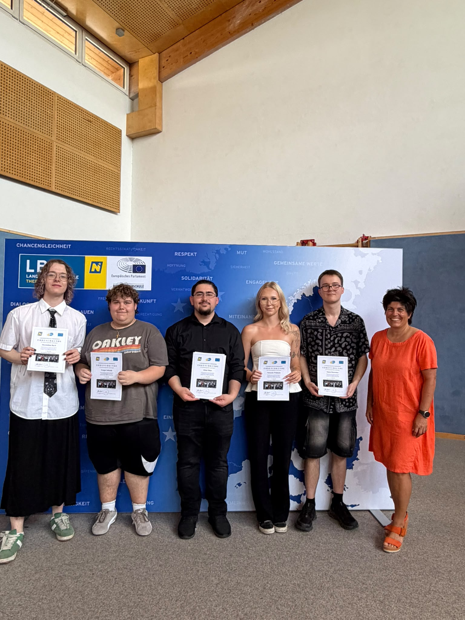 Abschlussfeier_The Road to Digital Freedom_21 Gruppenfoto der Klasse 3TKa, in der Turnhalle vor einer blau-weißen EU-Präsentationswand; einige halten Zertifikate oder Unterlagen in der Hand, rechts steht eine Lehrperson ( Eine Gruppe Jugendlicher steht in der Turnhalle vor einer blau-weißen Präsentationswand; einige halten Zertifikate oder Unterlagen in der Hand.