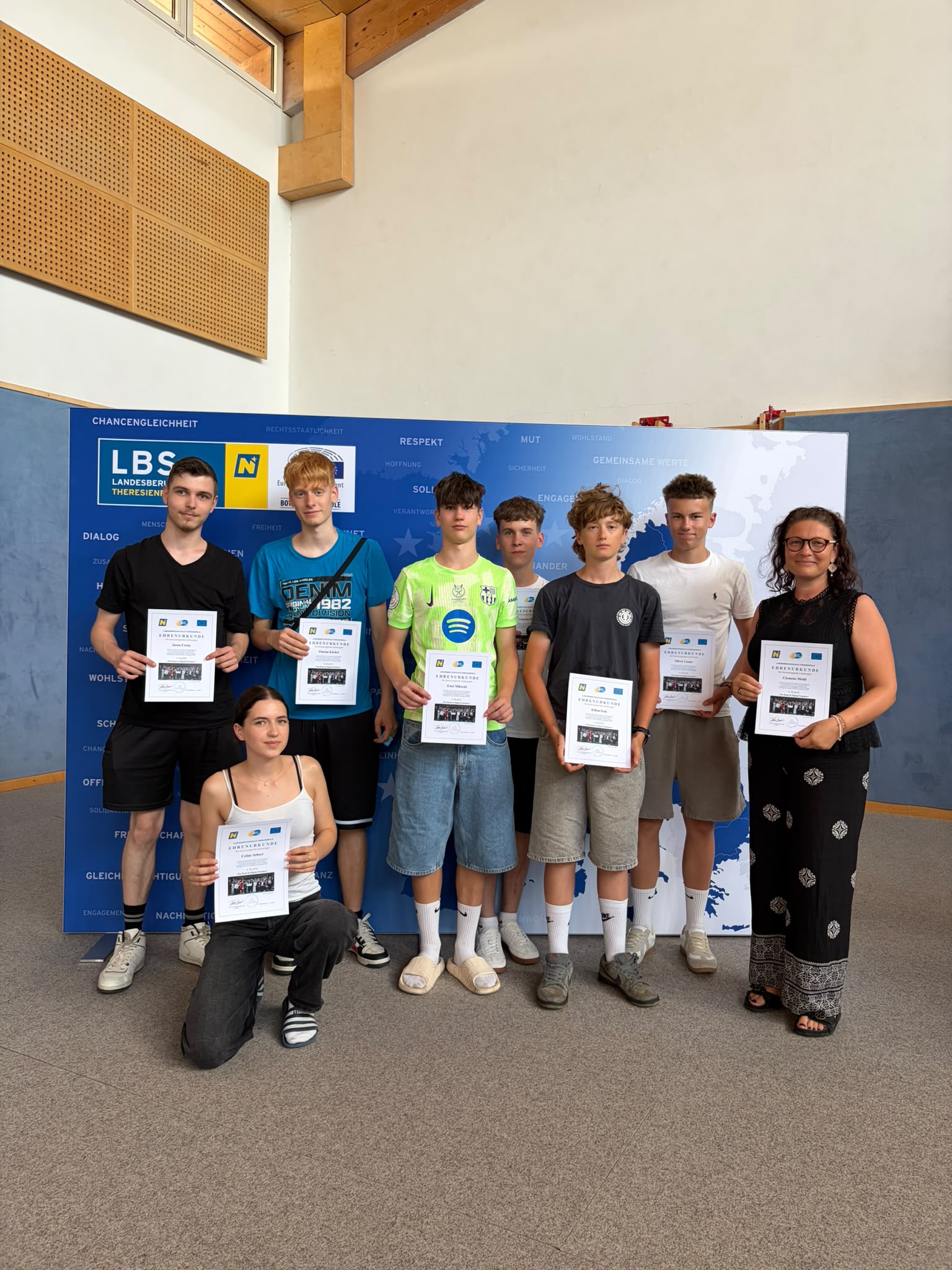 Abschlussfeier_The Road to Digital Freedom_20 Gruppenfoto der Klasse 1SPa, in der Turnhalle vor einer blau-weißen EU-Präsentationswand; einige halten Zertifikate oder Unterlagen in der Hand, rechts steht eine Lehrperson ( Eine Gruppe Jugendlicher steht in der Turnhalle vor einer blau-weißen Präsentationswand; einige halten Zertifikate oder Unterlagen in der Hand.
