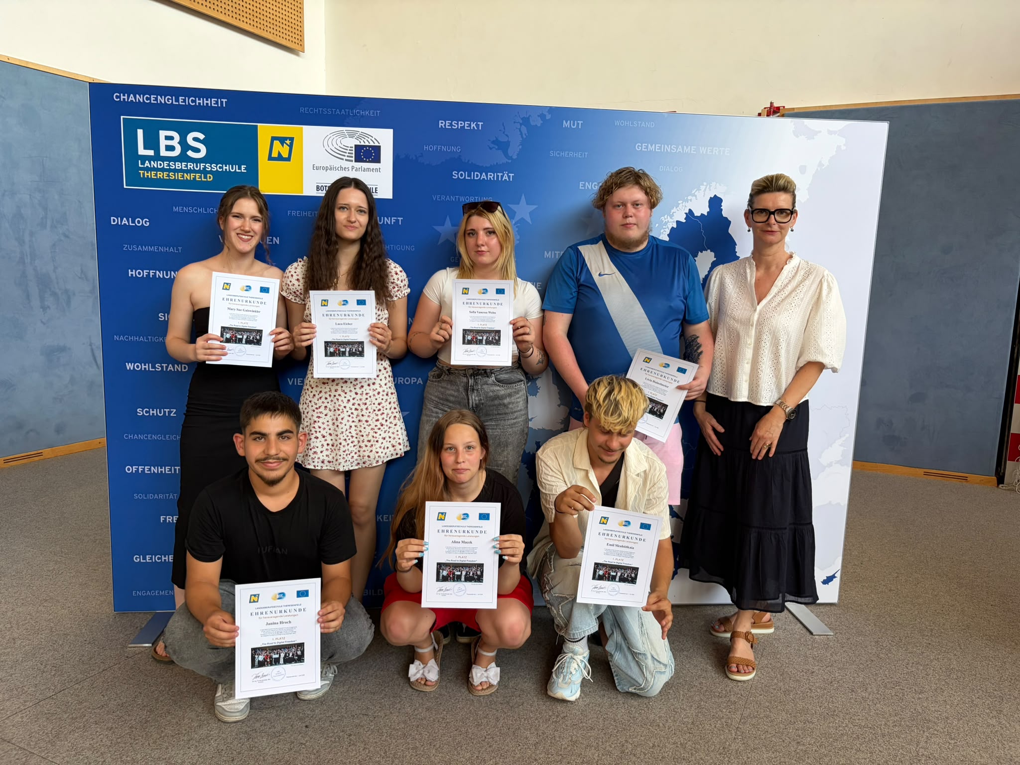 Abschlussfeier_The Road to Digital Freedom_18 Gruppenfoto der Klasse 2LH8a, in der Turnhalle vor einer blau-weißen EU-Präsentationswand; einige halten Zertifikate oder Unterlagen in der Hand, rechts steht eine Lehrperson ( Eine Gruppe Jugendlicher steht in der Turnhalle vor einer blau-weißen Präsentationswand; einige halten Zertifikate oder Unterlagen in der Hand.
