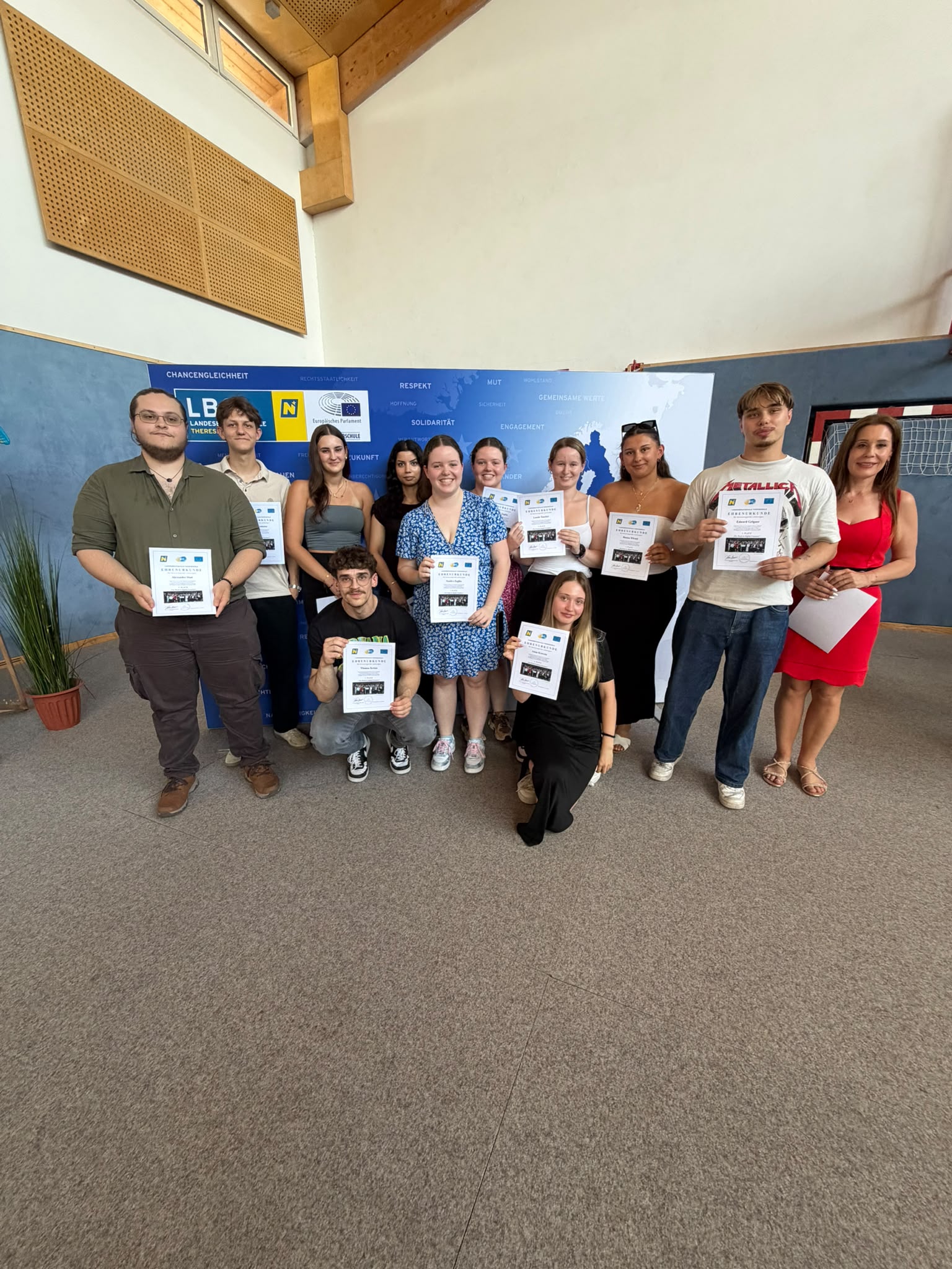 Abschlussfeier_The Road to Digital Freedom_16 Gruppenfoto der Klasse 3GH2b, in der Turnhalle vor einer blau-weißen EU-Präsentationswand; einige halten Zertifikate oder Unterlagen in der Hand, rechts steht eine Lehrperson ( Eine Gruppe Jugendlicher steht in der Turnhalle vor einer blau-weißen Präsentationswand; einige halten Zertifikate oder Unterlagen in der Hand.