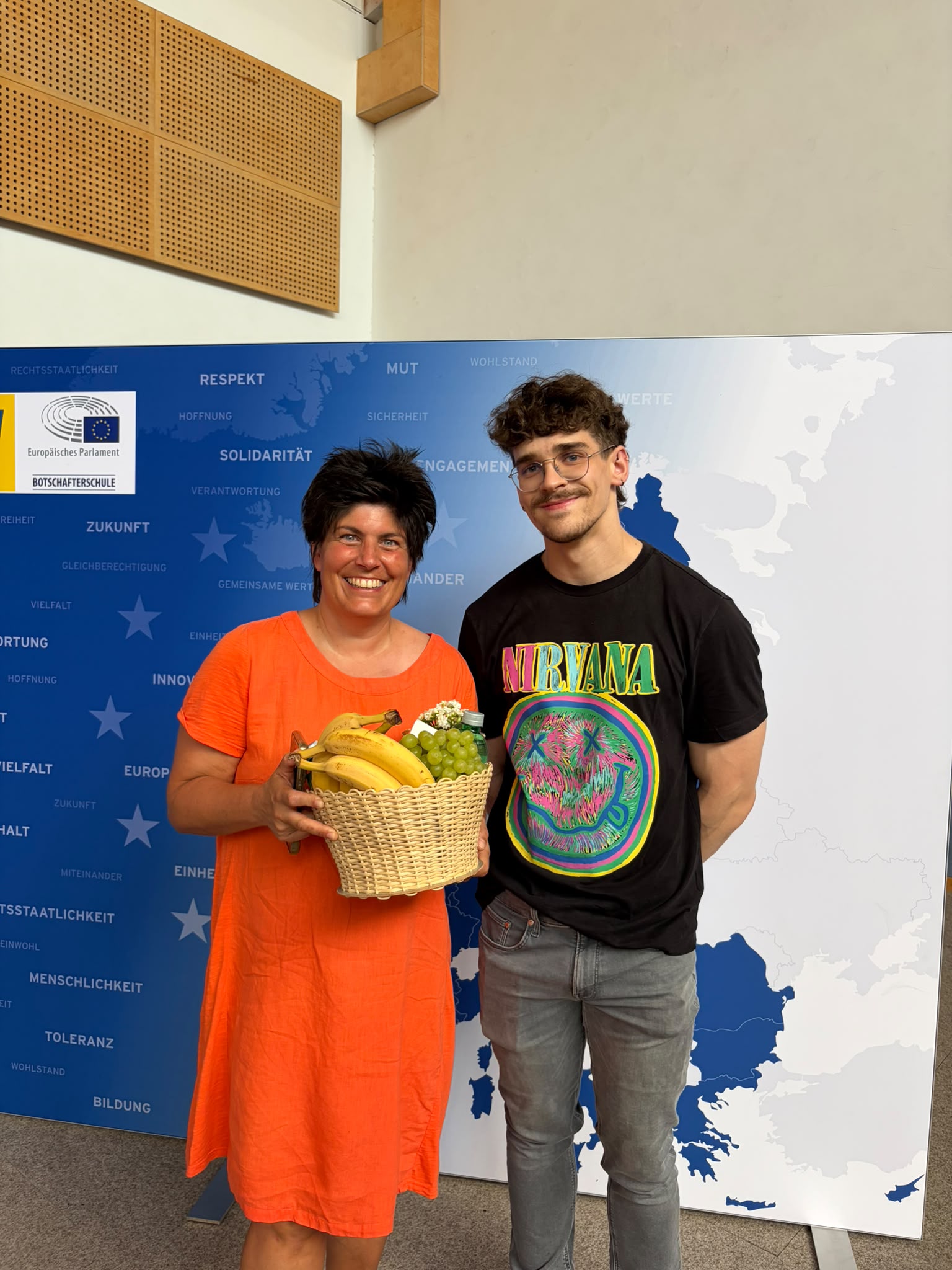 Abschlussfeier_The Road to Digital Freedom_15 Eine Lehrerin und ein Jugendlicher stehen vor einer blau-weißen EU-Präsentationswand; die Lehrerin hält einen Obstkorb, der Jugendliche steht daneben und lächelt.