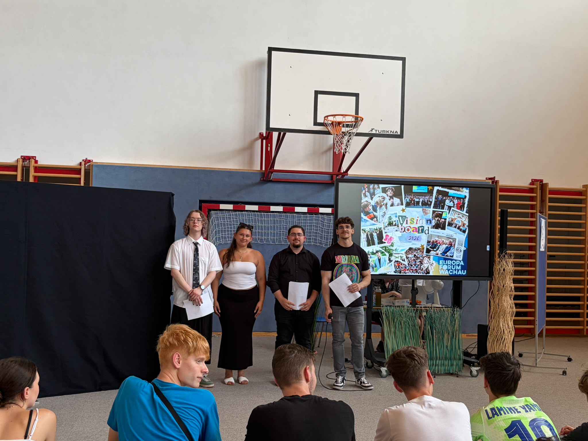 Abschlussfeier_The Road to Digital Freedom_13 Vier Schüler:innen stehen in einer Turnhalle vor einer verhüllten Präsentationswand und einem digitalen Screen, auf dem ein Visionboard mit verschiedenen Fotos zu sehen ist,m und halten Unterlagen in der Hand.