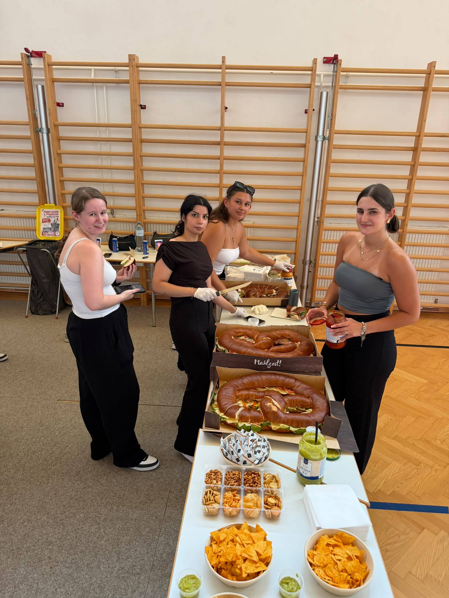 Abschlussfeier_The Road to Digital Freedom_3 Vier Schüler:innen servieren in der Turnhalle eine Jause mit belegten Brezen und Snacks.