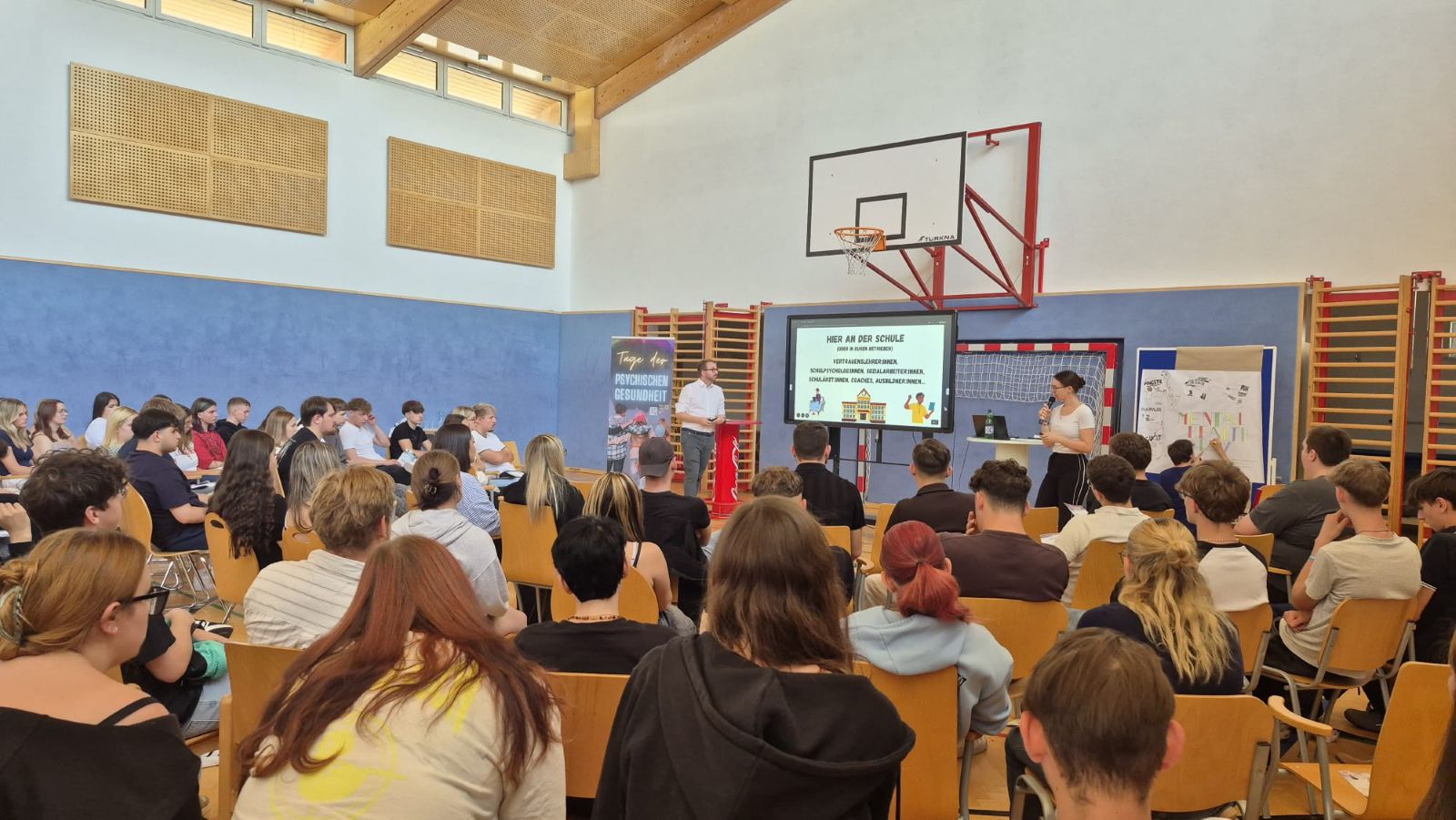 Mental Health Days 1.LG 2026/27 Schülerinnen und Schüler sitzen bei einer Veranstaltung in der Turnhalle und verfolgen einen Vortrag über psychische Gesundheit.