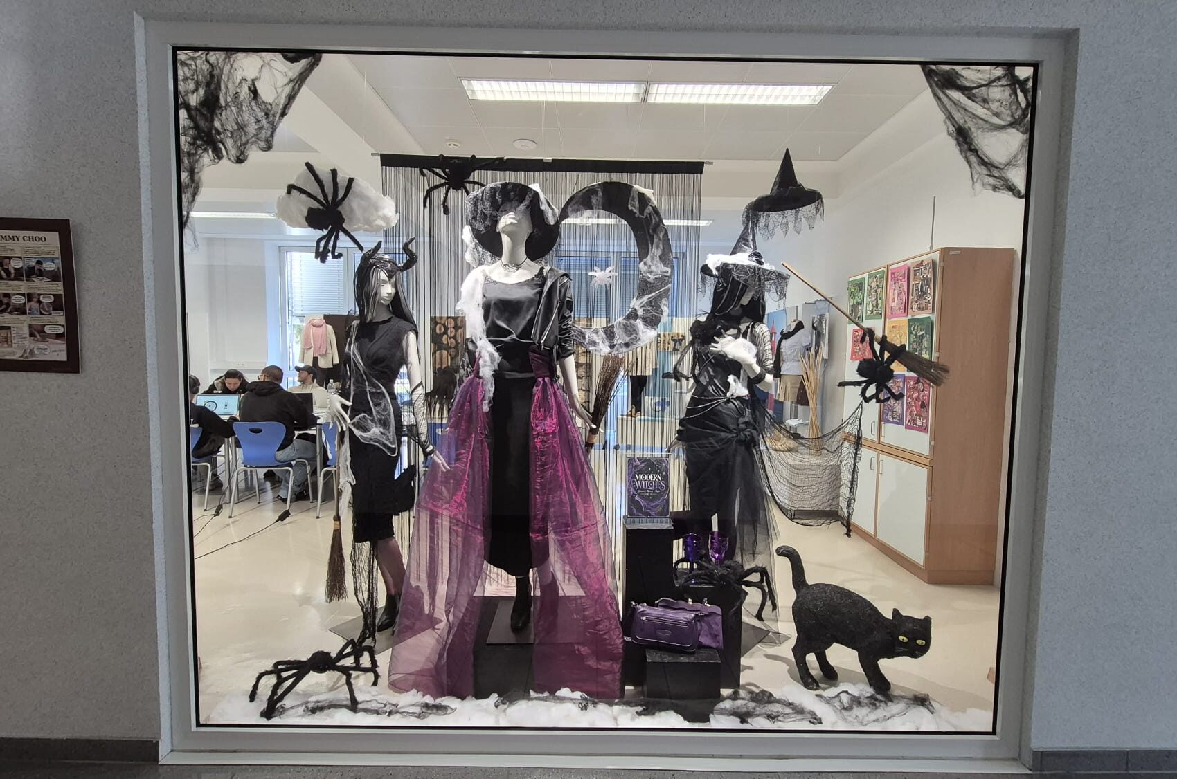 Halloween 2026 Halloween-Schaufenster mit drei Schaufensterpuppen in schwarzer Damenmode, violetten Accessoires, Hexenhüten, Spinnweben, Besen und einer schwarzen Katze.